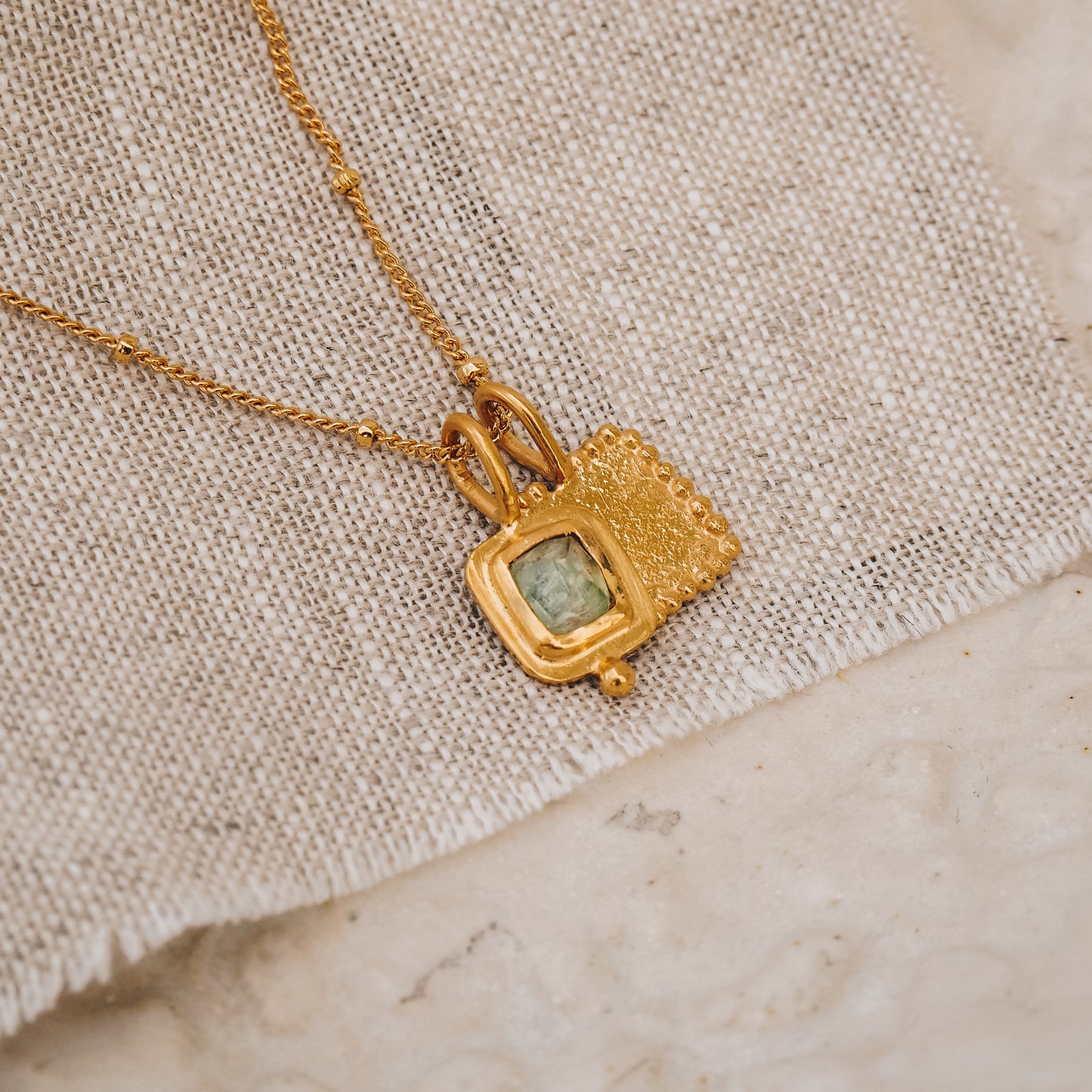 Two dainty, square, gold pendants, one with a rose cut blue tourmaline, suspended from a fine chain 
