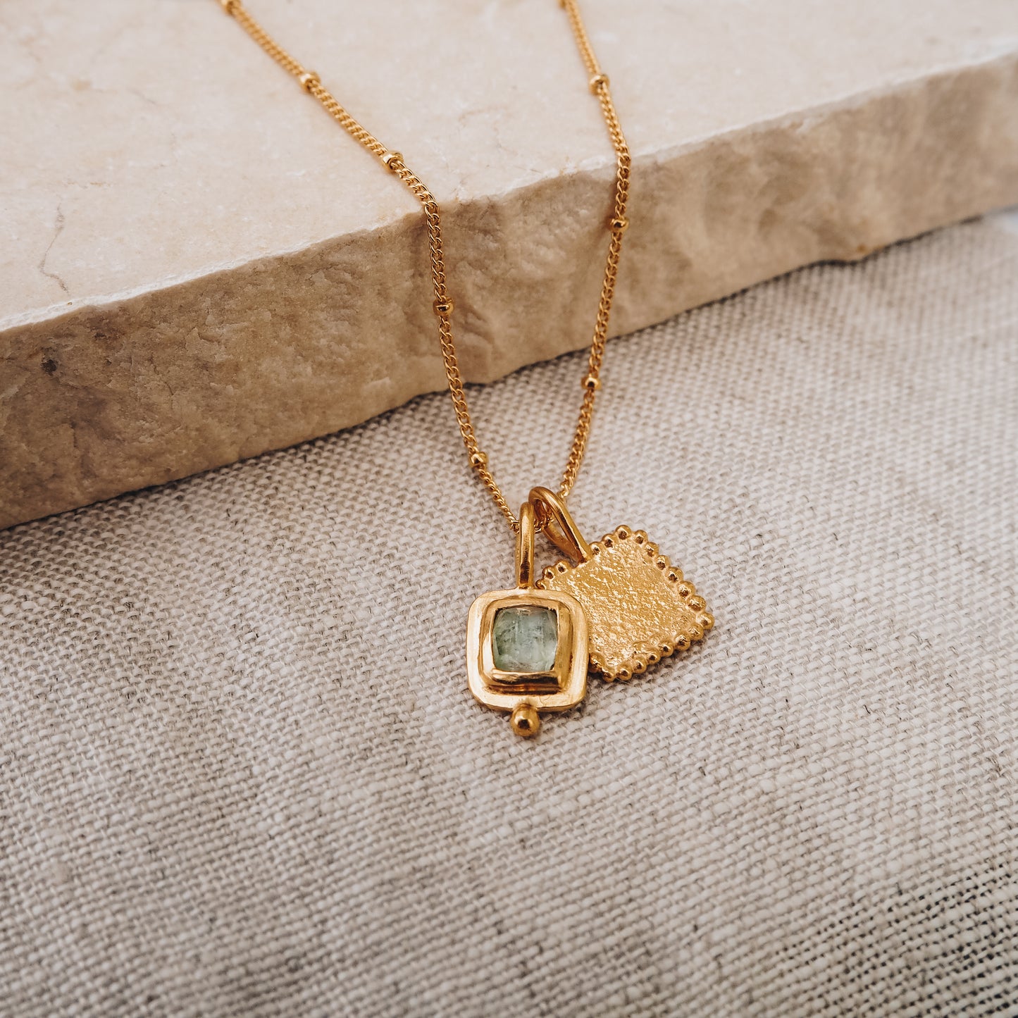 A gold satellite chain with an organically textured pendant and a blue tourmaline gemstone.