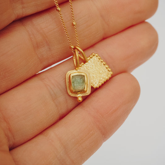 A small gold necklace with a pair of square pendants, one textured and one set with a blue rose cut tourmaline
