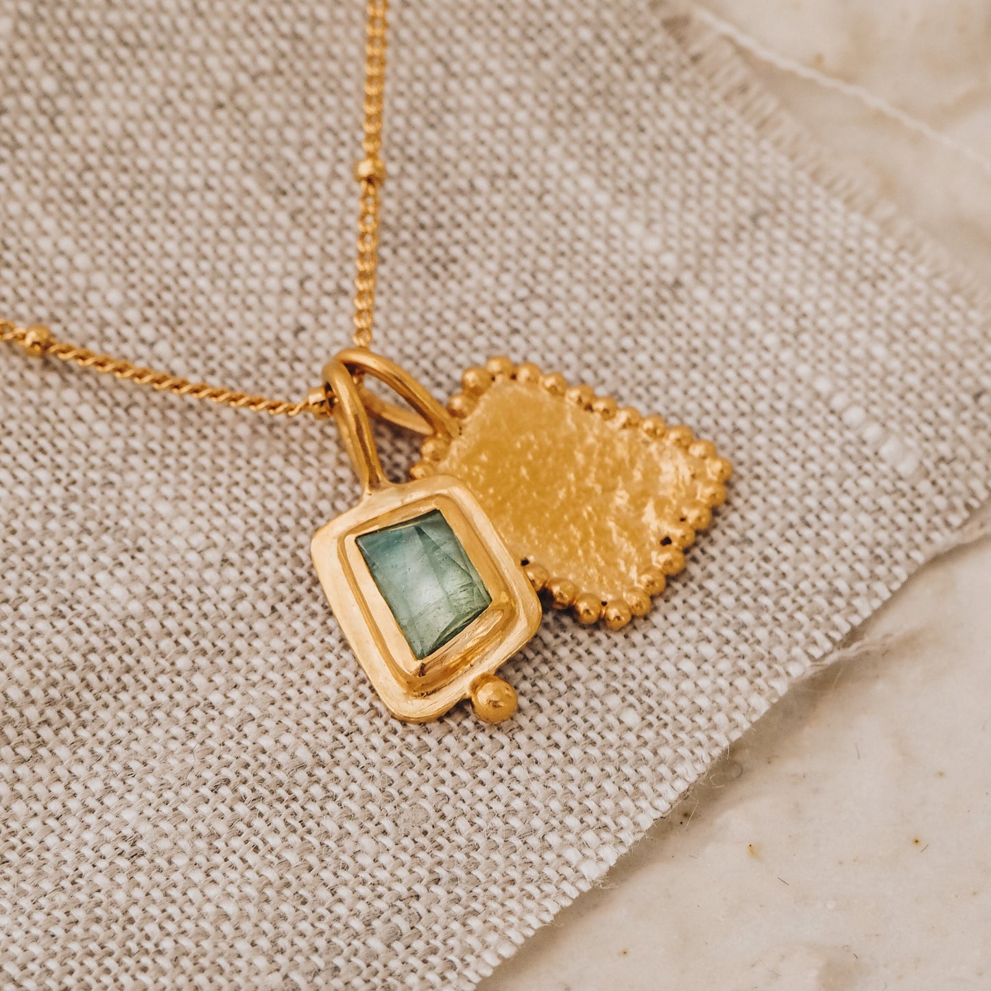 Square gold pendant with a vibrant blue rose cut tourmaline gemstone and meticulous granulation detail, elegantly suspended from a delicate satellite chain.