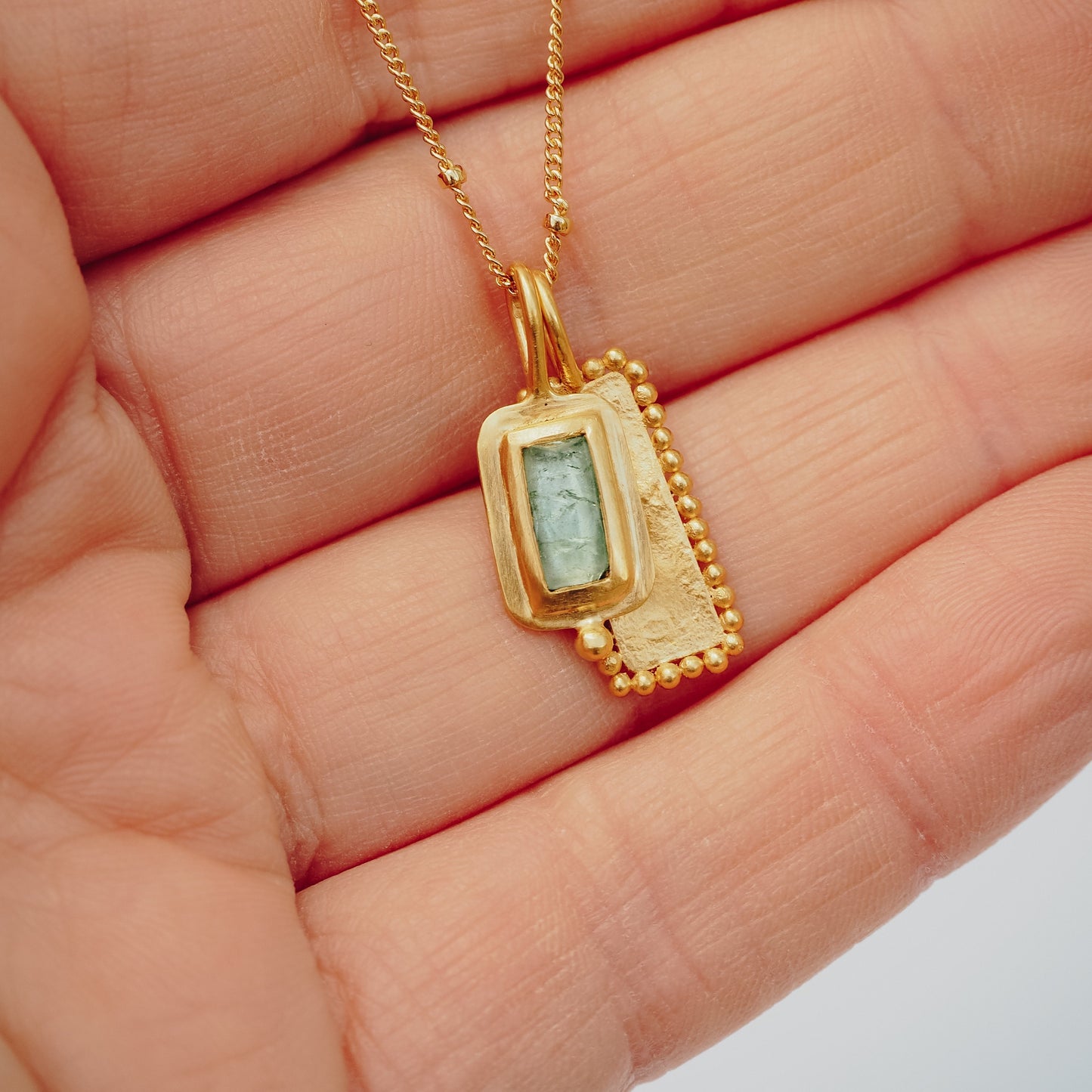 Exquisite square gold pendant adorned with a vibrant blue rose cut tourmaline gemstone and intricate granulation, hanging delicately from a satellite chain.