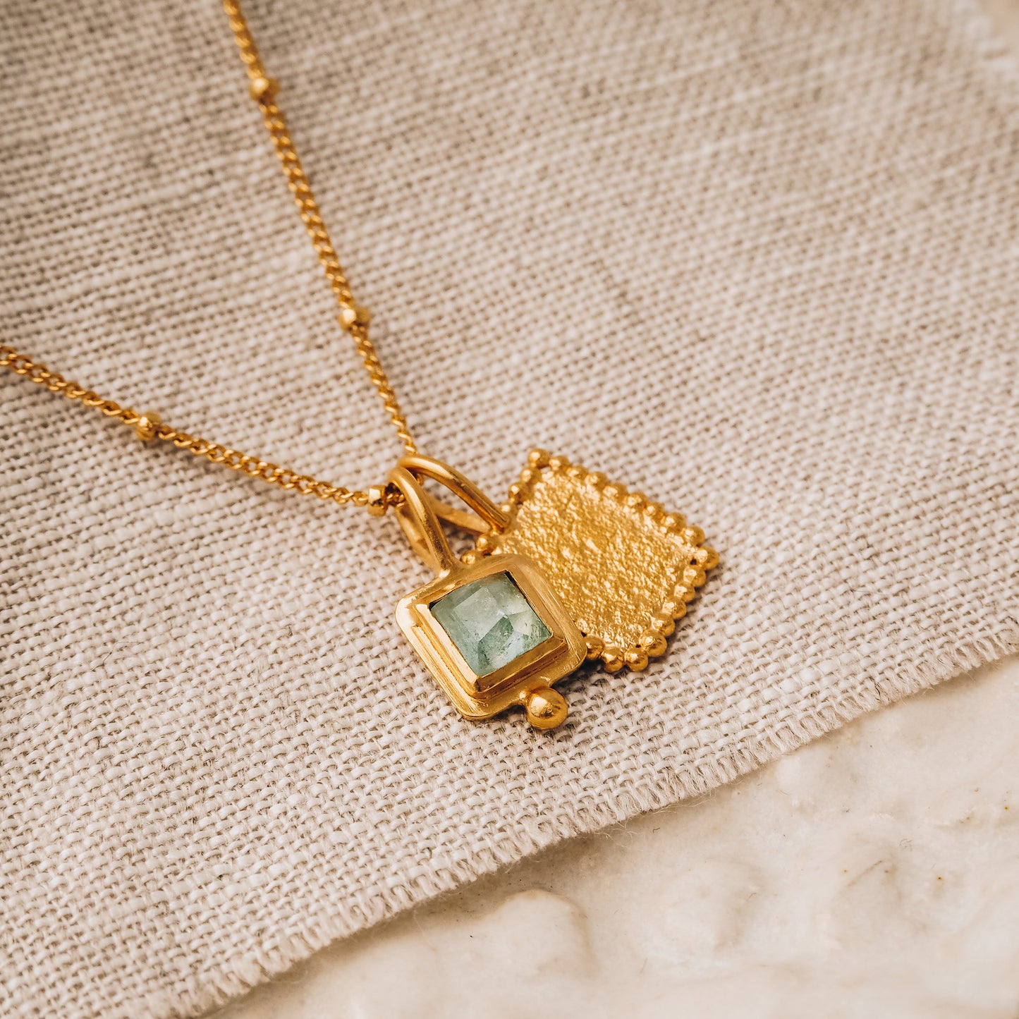 Square gold pendant adorned with a vibrant blue rose cut tourmaline gemstone and exquisite granulation, hanging gracefully from a delicate satellite chain.