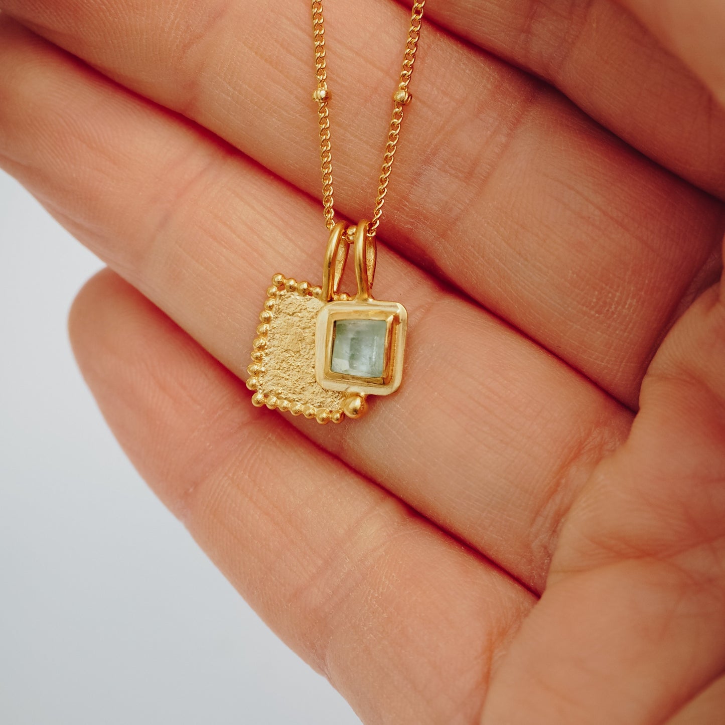 Square gold pendant adorned with a vibrant blue rose cut tourmaline gemstone and meticulous granulation work, hanging delicately from a satellite chain.
