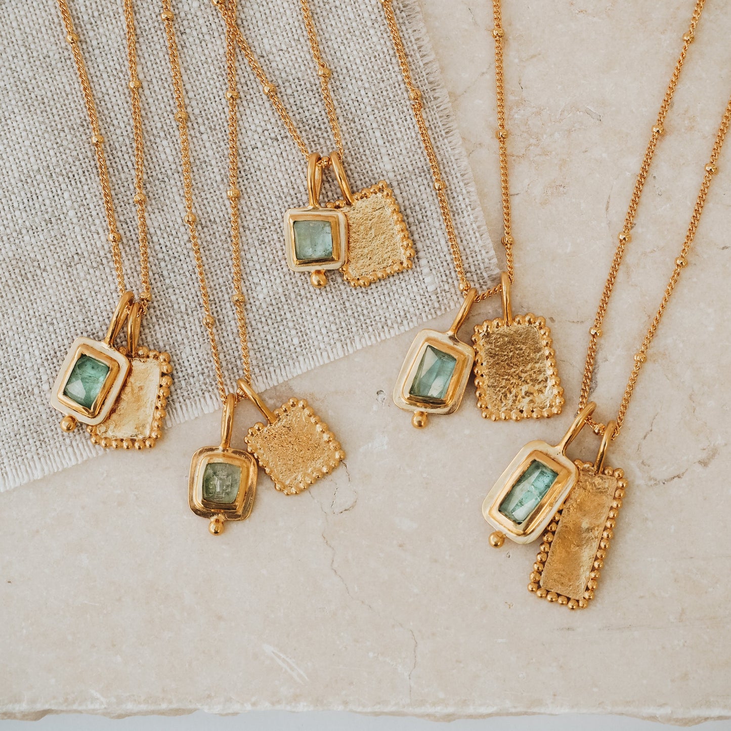 Collection of elegant rich gold necklaces, each featuring a square pendant adorned with a mesmerizing ocean blue rose cut tourmaline gemstone, textured surfaces, and meticulous granulation work