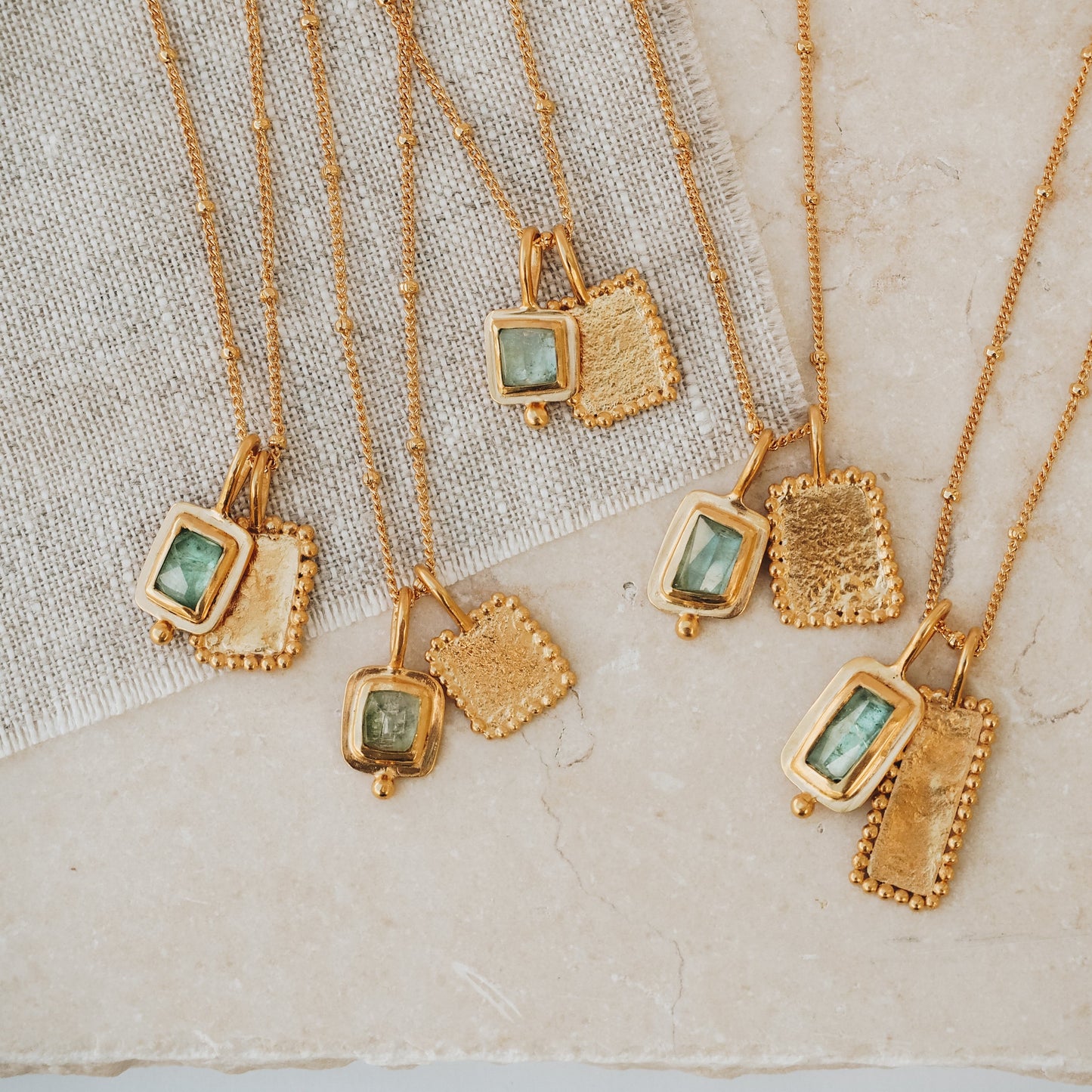 Collection of handcrafted rich gold necklaces with square pendants, adorned with ocean blue rose cut tourmaline gemstones, textured surfaces, and intricate granulation detailing.