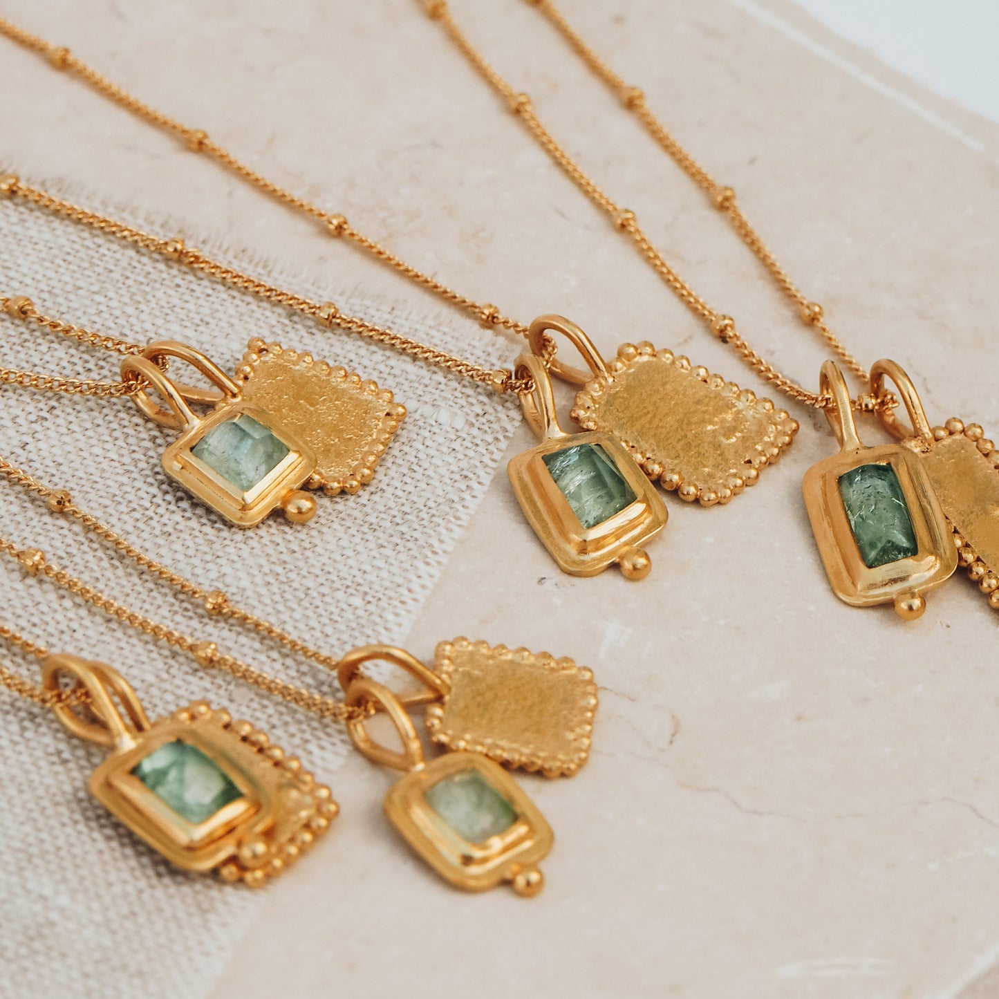 Group of exquisite rich gold necklaces featuring square pendants, captivating ocean blue rose cut tourmaline gemstones, textured surfaces, and meticulous granulation work.