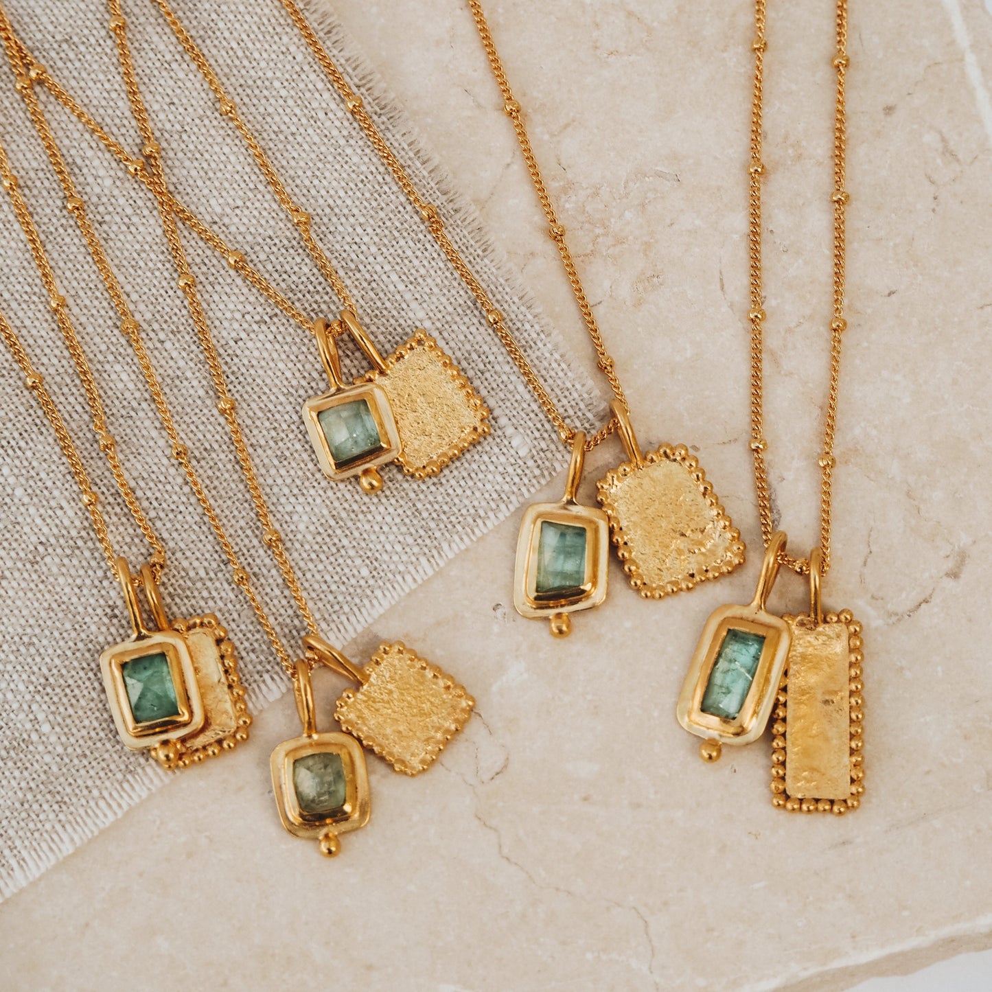 Collection of artisan-crafted rich gold necklaces, each with a square pendant embellished with an enchanting ocean blue rose cut tourmaline gemstone, textured surfaces, and delicate granulation accents.